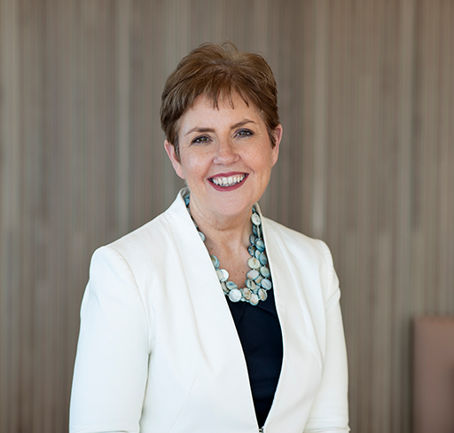 A person wearing a white blazer, dark top, and a light blue beaded necklace stands in front of a softly lit wooden panel background.