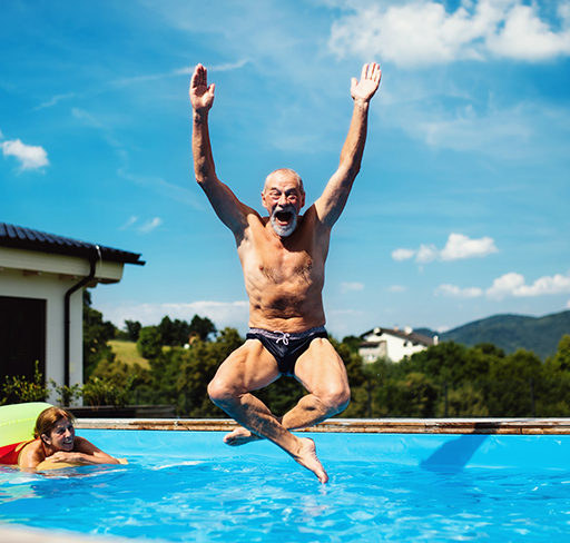 An elderly man joyfully jumps into a swimming pool with his arms raised, while a woman watches from the poolside, set against a backdrop of a sunny day and rolling hills.