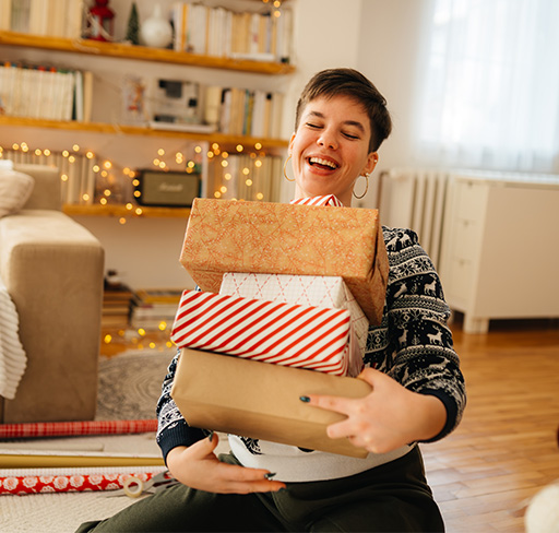 A person wearing a winter sweater sits in a cosy living room holding a stack of wrapped holiday gifts, with fairy lights and bookshelves in the background