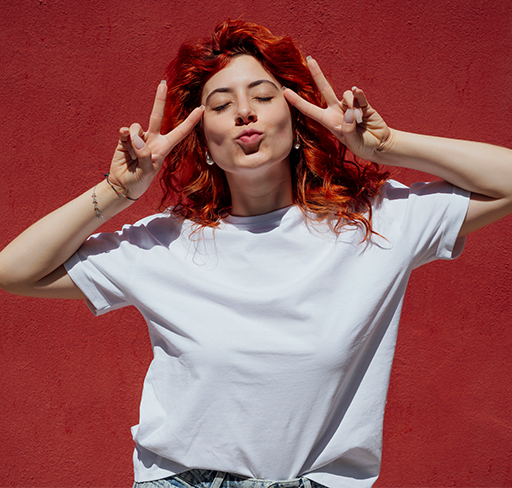 A person with vibrant red hair poses in front of a bright red wall, making a playful gesture with both hands forming peace signs. They are wearing a plain white t-shirt, standing out against the bold background.