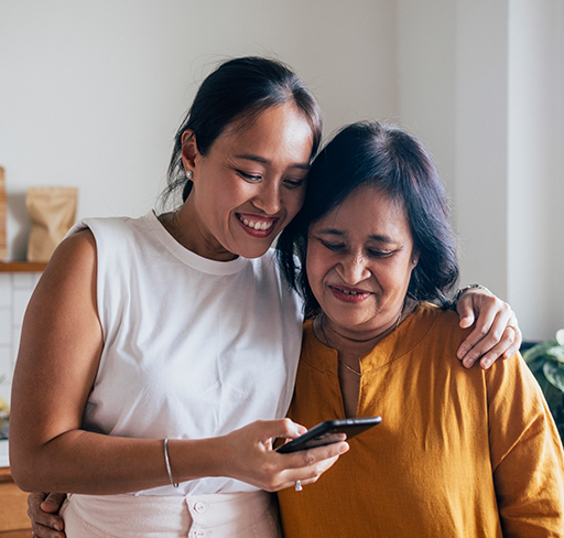 A mother and daughter standing close together in a kitchen, one with their arm around the other, both looking at a mobile phone.