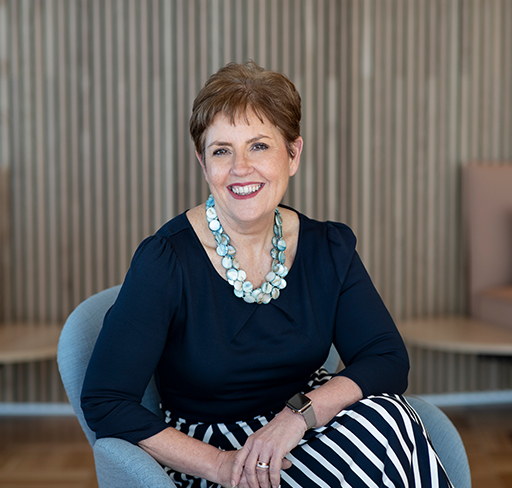 A woman with short brown hair sits in a modern chair, wearing a navy top, a chunky light blue necklace, and a navy-and-white striped skirt, with a wooden slat wall in the background.