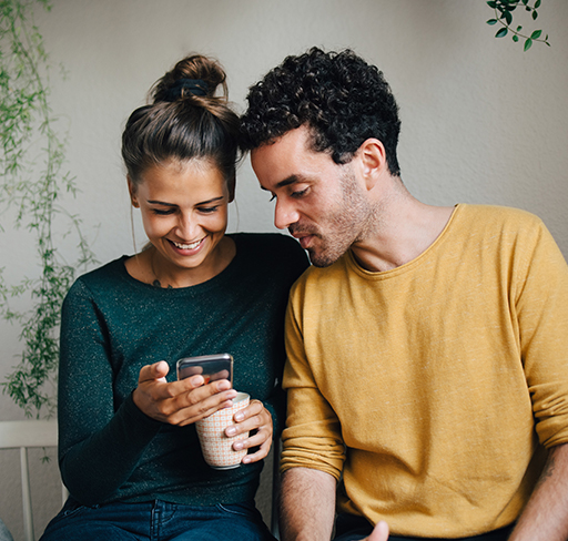 A woman and a man sit closely together on a bench, with the woman holding a smartphone and a drink while the man leans in to look at the screen.