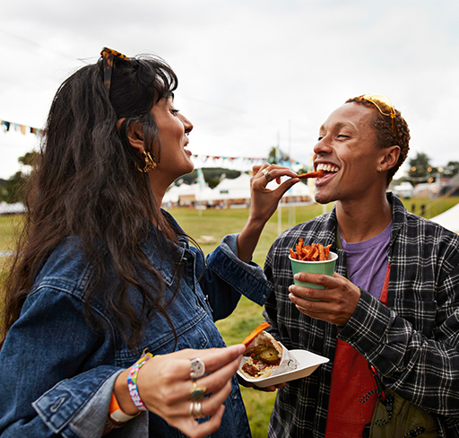 Two people are outdoors at a festival or event, with one holding a cup of fries and the other reaching for a fry.
