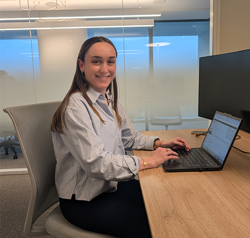 A person with long brown hair, wearing a light blue striped shirt and dark trousers, sits at a desk in a modern office space and types on a laptop, with a large monitor and glass walls in the background.