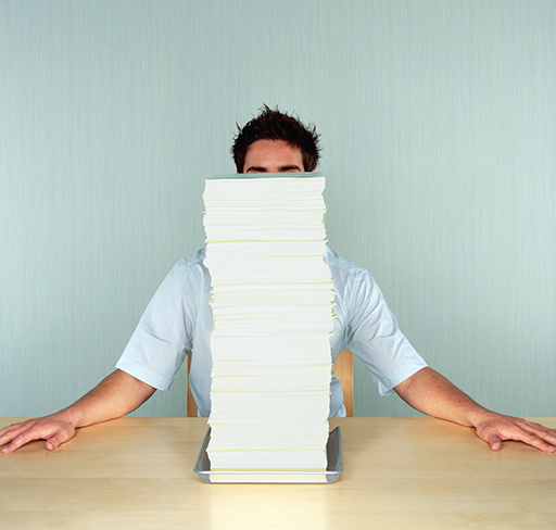 A person sits at a desk with their arms resting on the surface, almost completely hidden behind a tall stack of white papers against a pale blue background.
