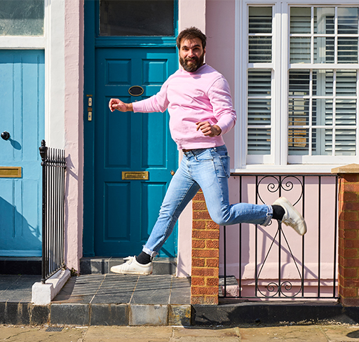 A person wearing a pink sweatshirt, blue jeans, and white sneakers is mid-jump on a footpath in front of colourful houses with blue doors and white-trimmed windows.