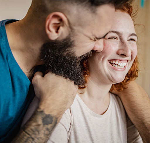 A man with a beard and tattooed arm leans closely towards another person, creating an intimate and affectionate moment.