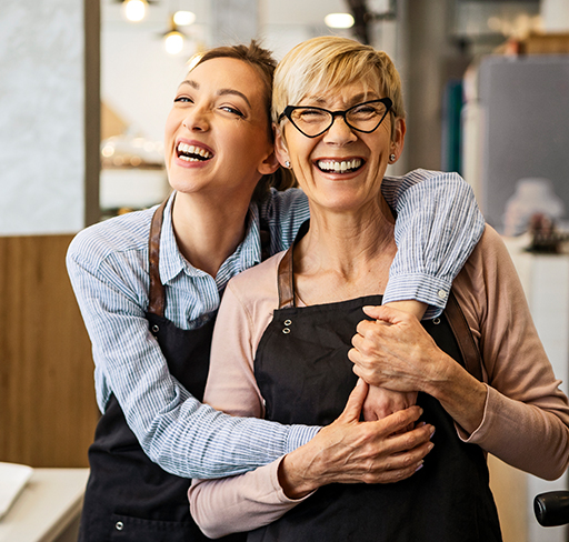 Two people wearing aprons are warmly embracing in a cozy kitchen setting, with soft lighting and wooden accents enhancing the inviting atmosphere.