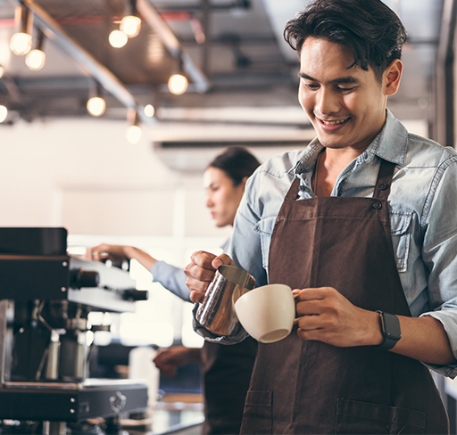 A barista in a brown apron pours milk into a cup in a warmly lit café.