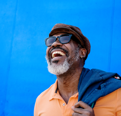 A man wearing an orange collared shirt sunglasses and a brown flat cap stands against a bright blue background, holding a blue jacket slung over their shoulder.
