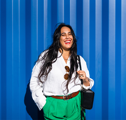 A woman stands against a bright blue corrugated wall, wearing a white blouse tucked into green trousers with a brown belt, sunglasses hanging at the neckline, and carrying a small black shoulder bag.