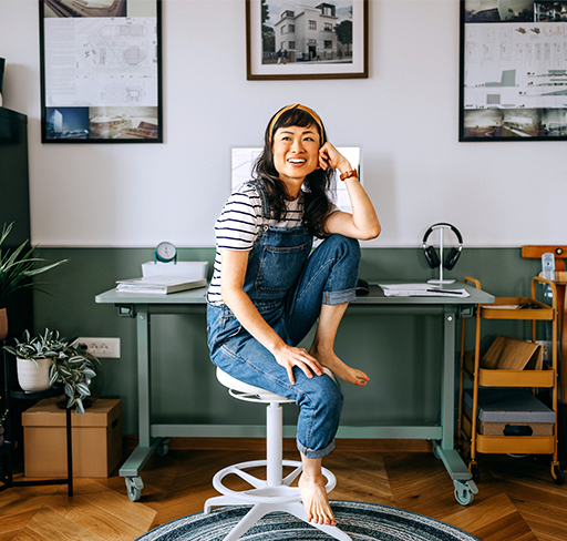 A person wearing denim overalls and a striped shirt is seated on a white swivel chair in a home office. The room features a green and white color scheme, with a desk in the background. 