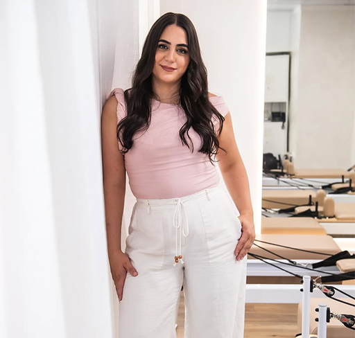 Woman in pink top and white pants stands inside a Pilates studio with reformers in the background